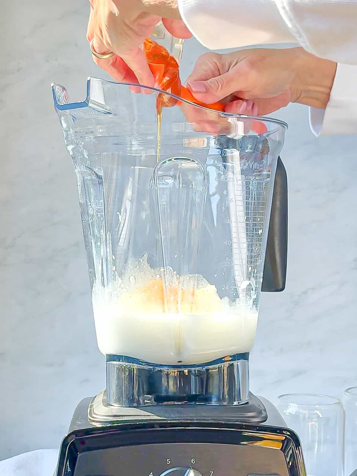 Hands pouring honey into a blender containing milk, poised to create a delicious mango yogurt smoothie on a light marble background.