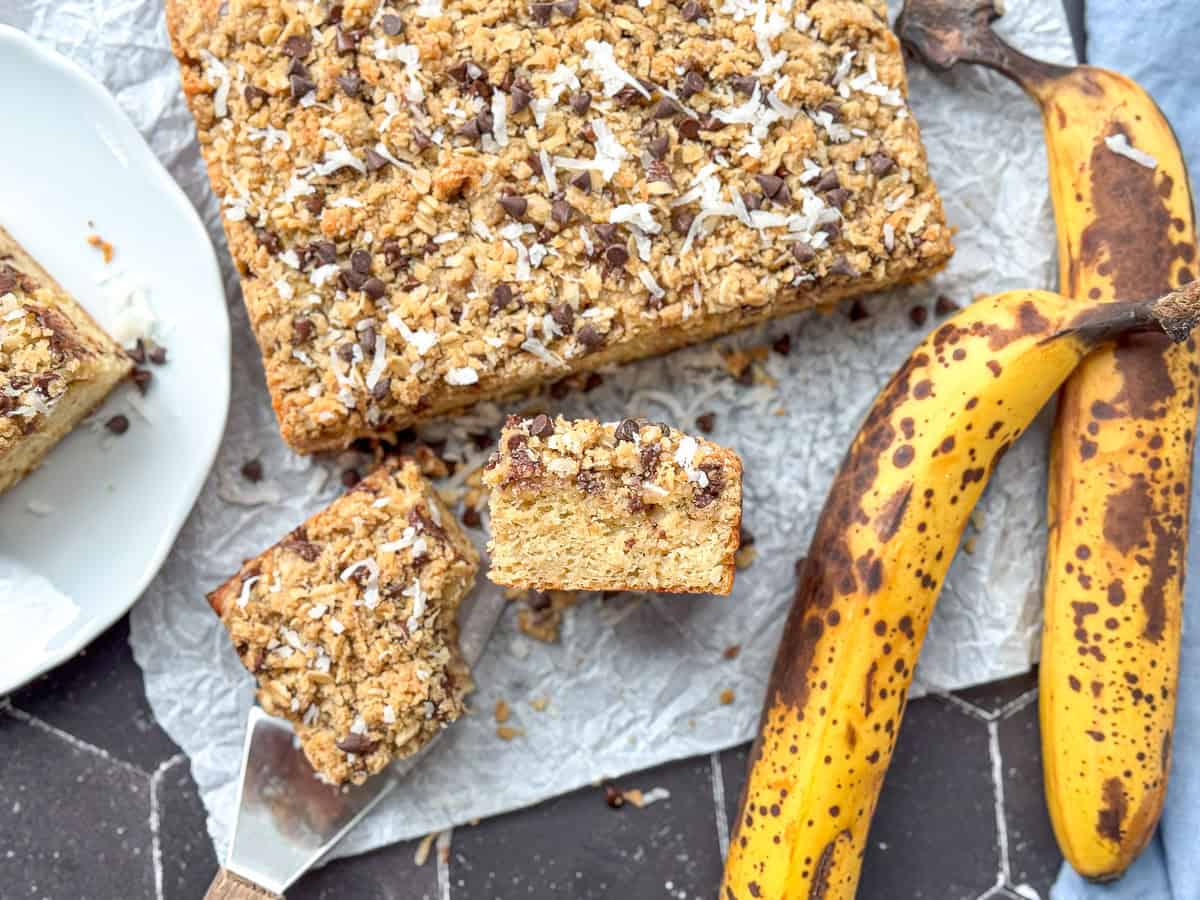 A slice of banana breakfast cake, with a bite taken out, on parchment paper along with the rest of the cake, a spatula, and two bananas.