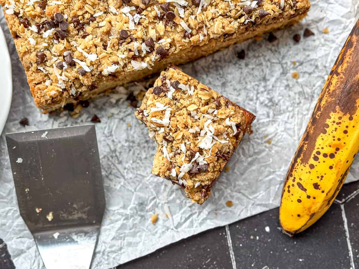 A slice of banana breakfast cake, with a bite taken out, on parchment paper along with the rest of the cake, a spatula, and a banana.