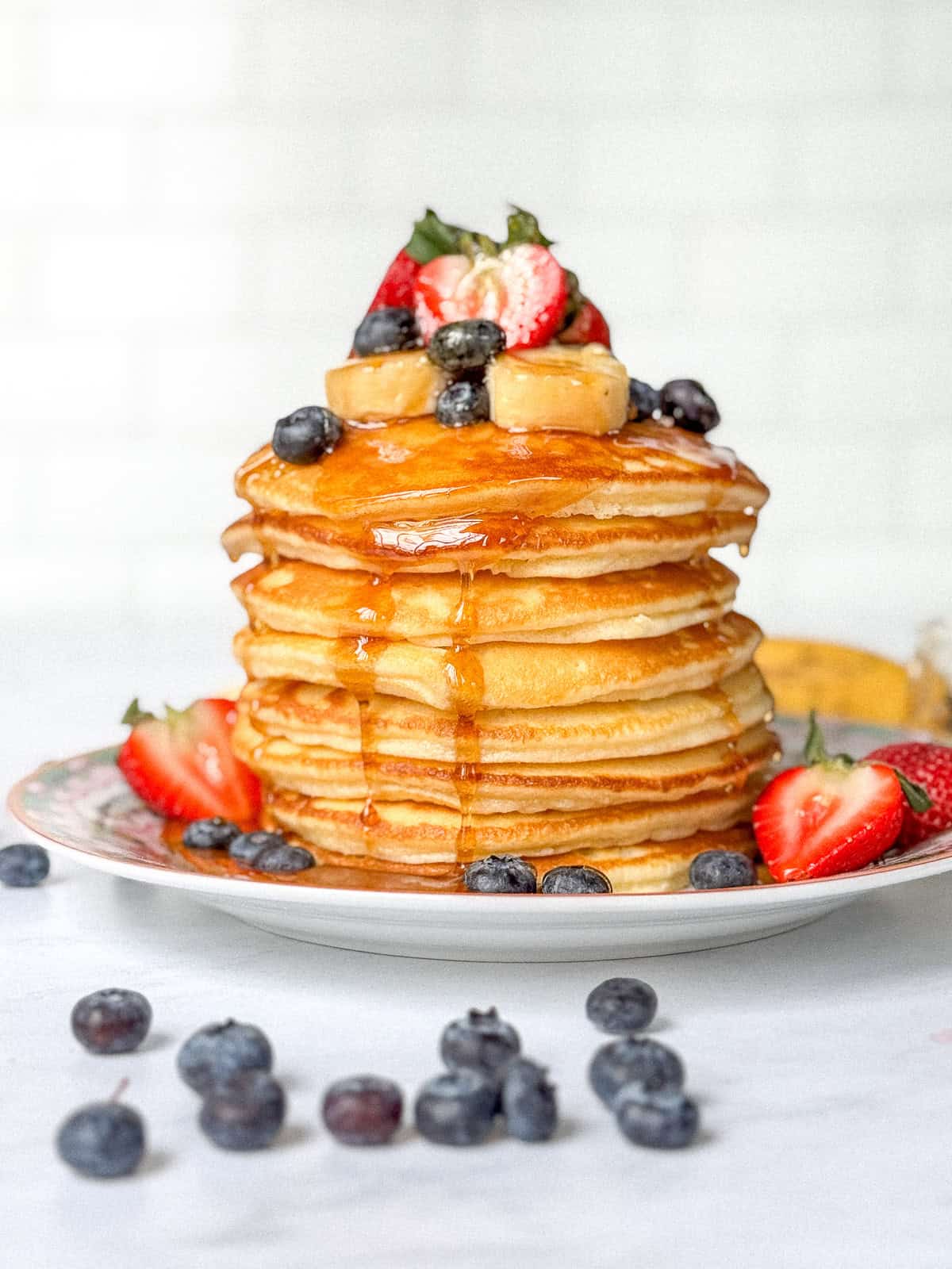 A stack of condensed milk pancakes dripping with maple syrup and with fruit on top and around the plate.