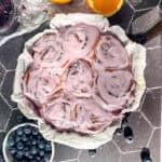 Image of a pan of blueberry rolls with blueberry frosting with oranges and blueberries in the background.
