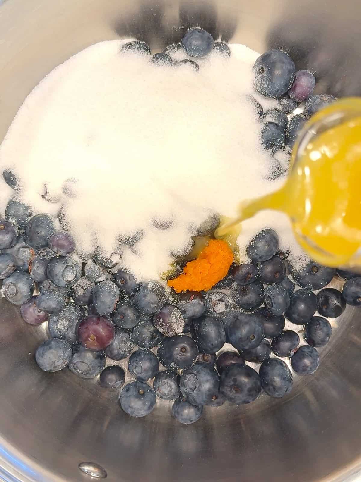 Pouring orange juice into a small pan containing blueberries, granulated sugar, and orange zest.