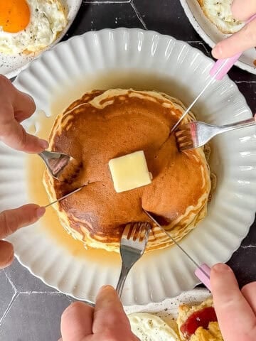 Three pairs of hands reaching in to cut up a short stack of pancakes.
