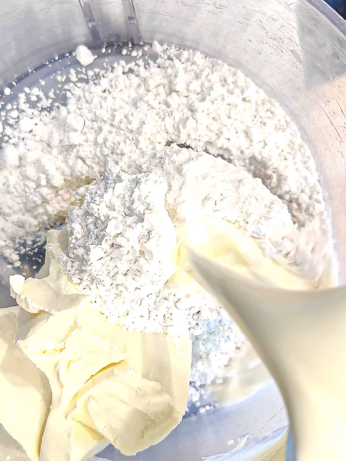 Adding heavy cream to powdered sugar and cream cheese in a food processor.