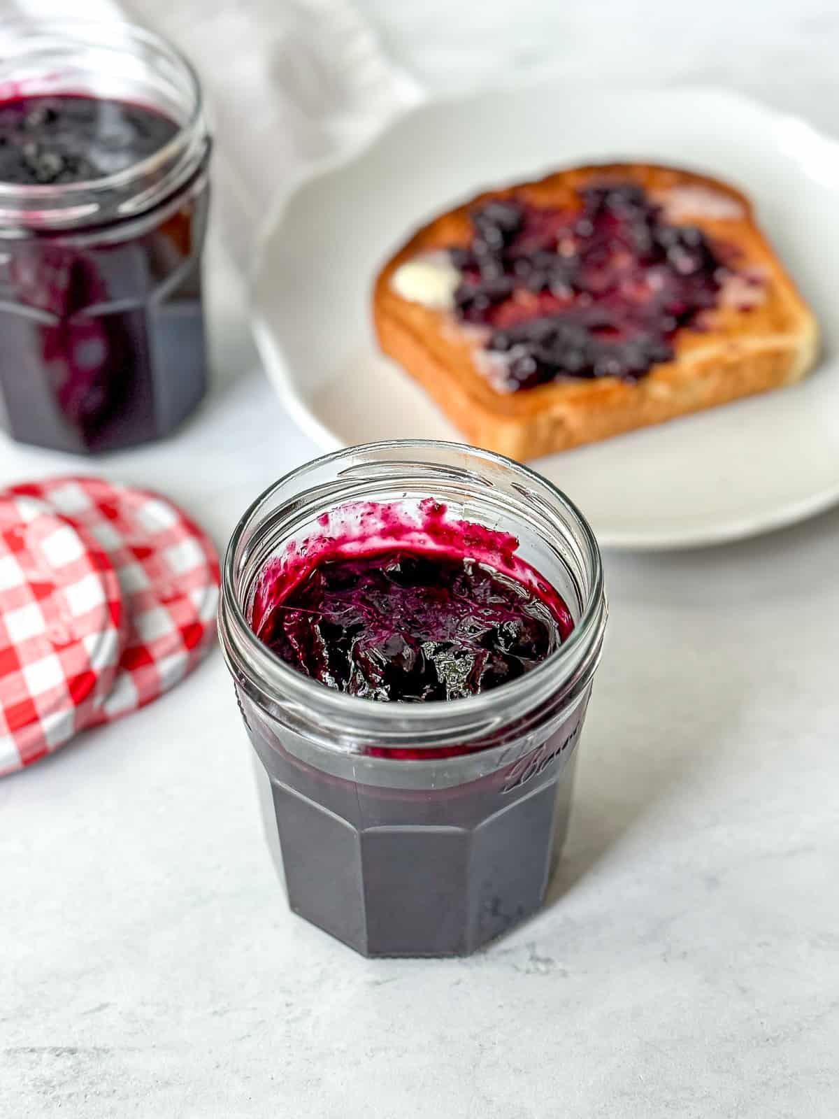 Two jars of blueberry jam and a slice of toast with jam on a white counter.