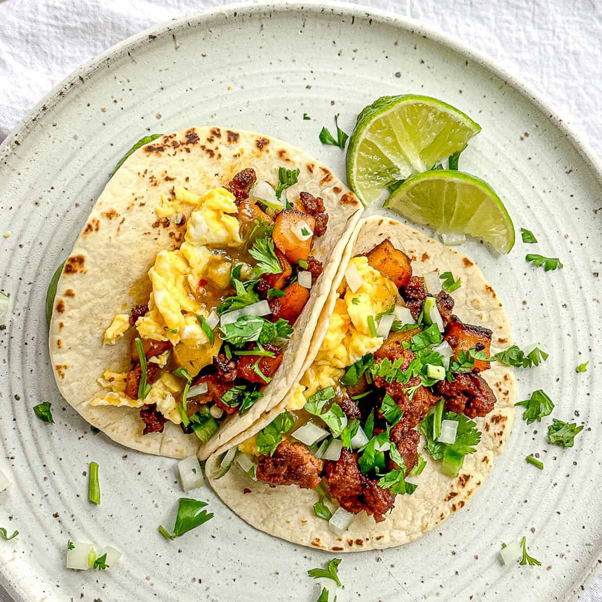 Two chorizo and potato breakfast tacos on a white plate with lime wedges.