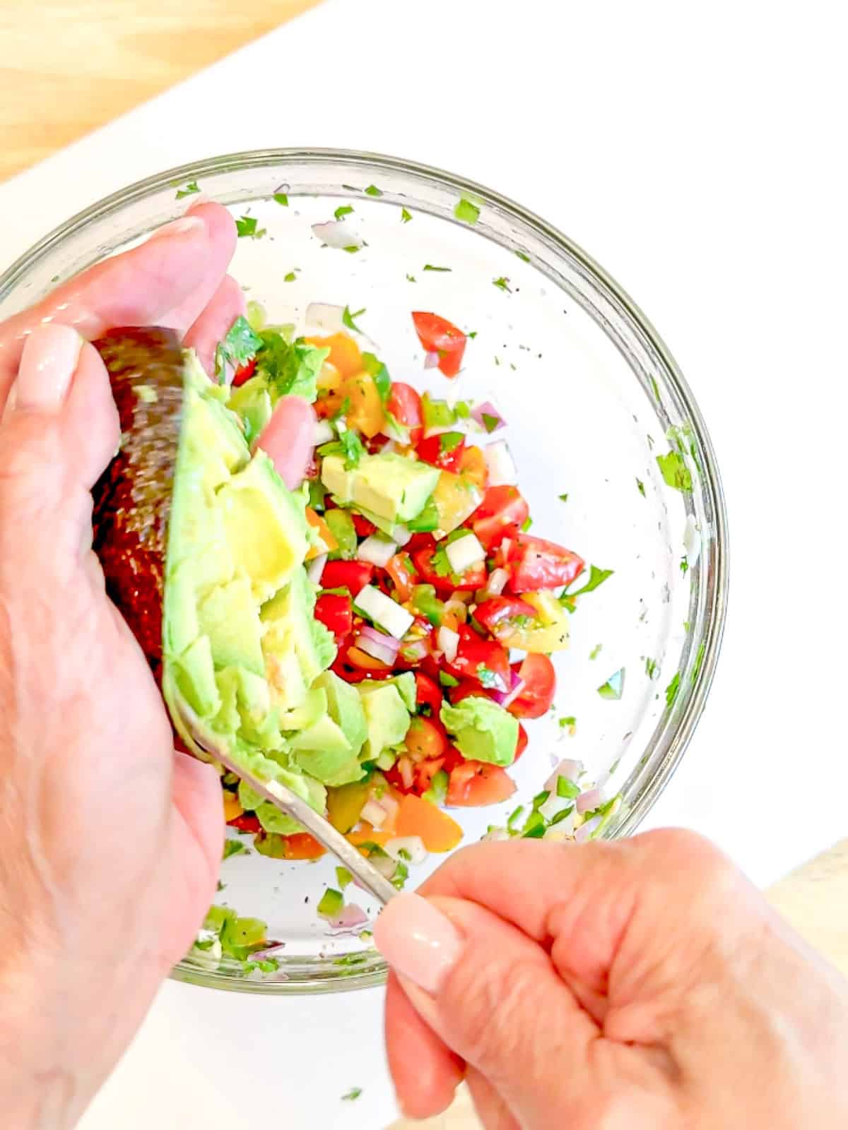 Adding avocado to pico de gallo in a glass bowl.