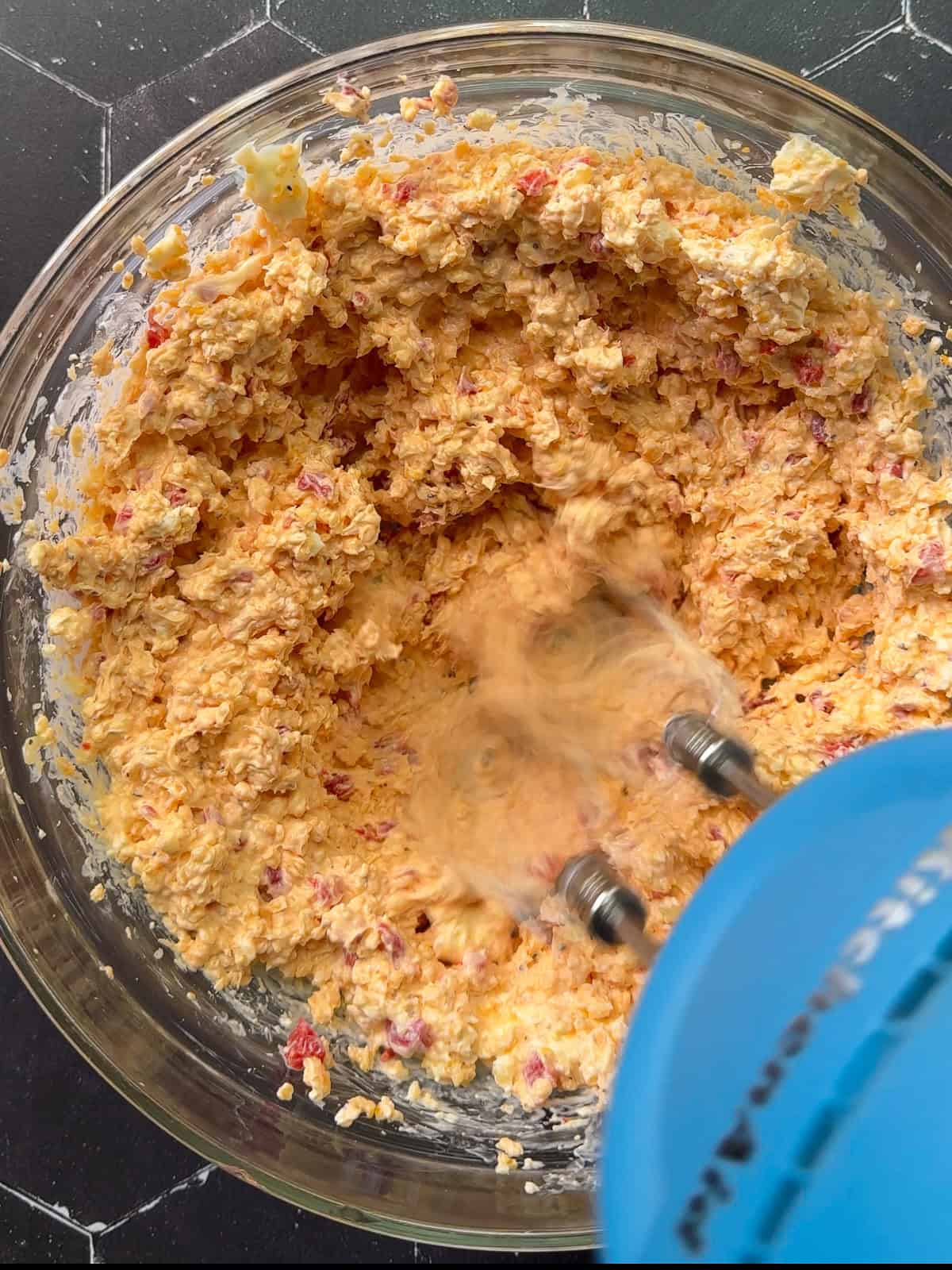 Using a hand mixer to mix pimento cheese in a glass bowl.