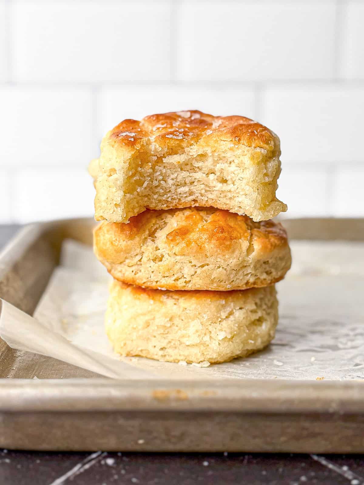 A stack of biscuits on a parchment paper lined sheet pan, the top one with a bite taken out.