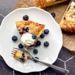 A slice of yogurt cake with blueberries on a white plate with the entire cake behind it on a black background.