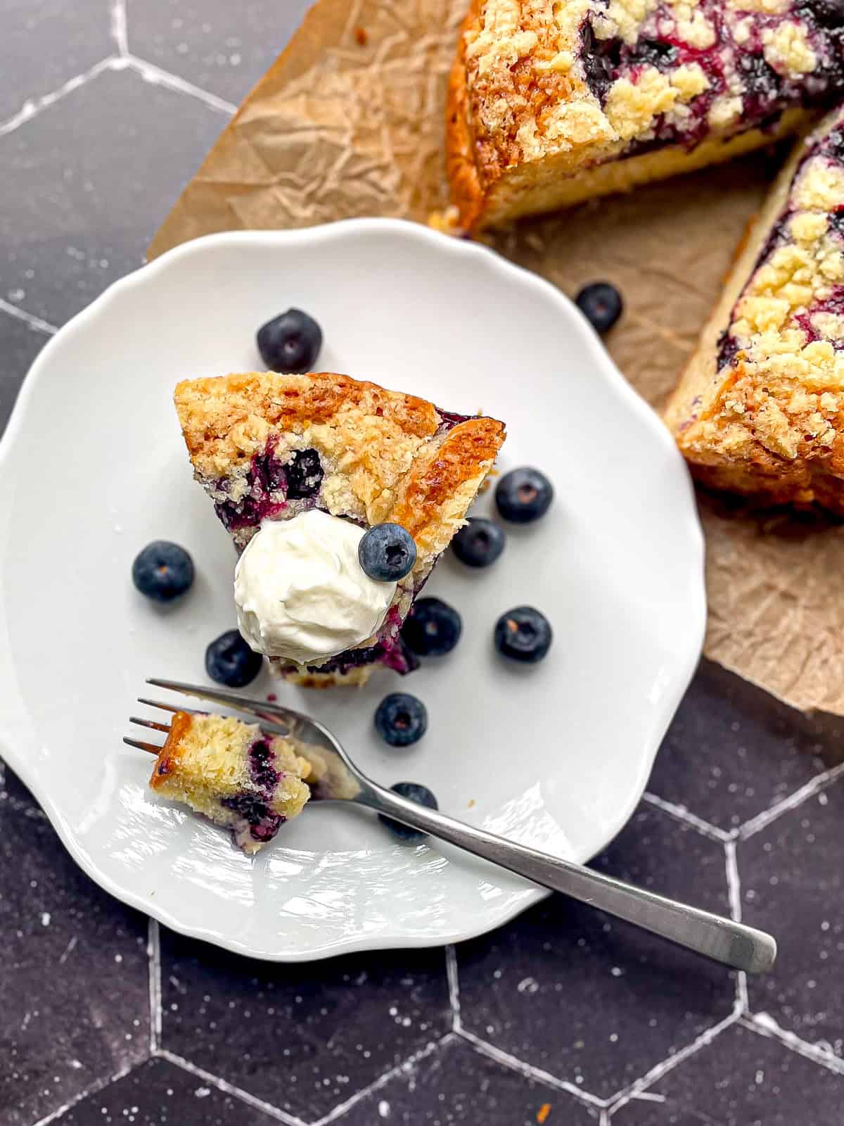 A slice of yogurt cake with blueberries on a white plate with the entire cake behind it on a black background.