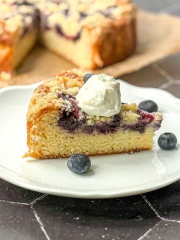 A slice of yogurt cake with blueberries on a white plate with the entire cake behind it on a black background.