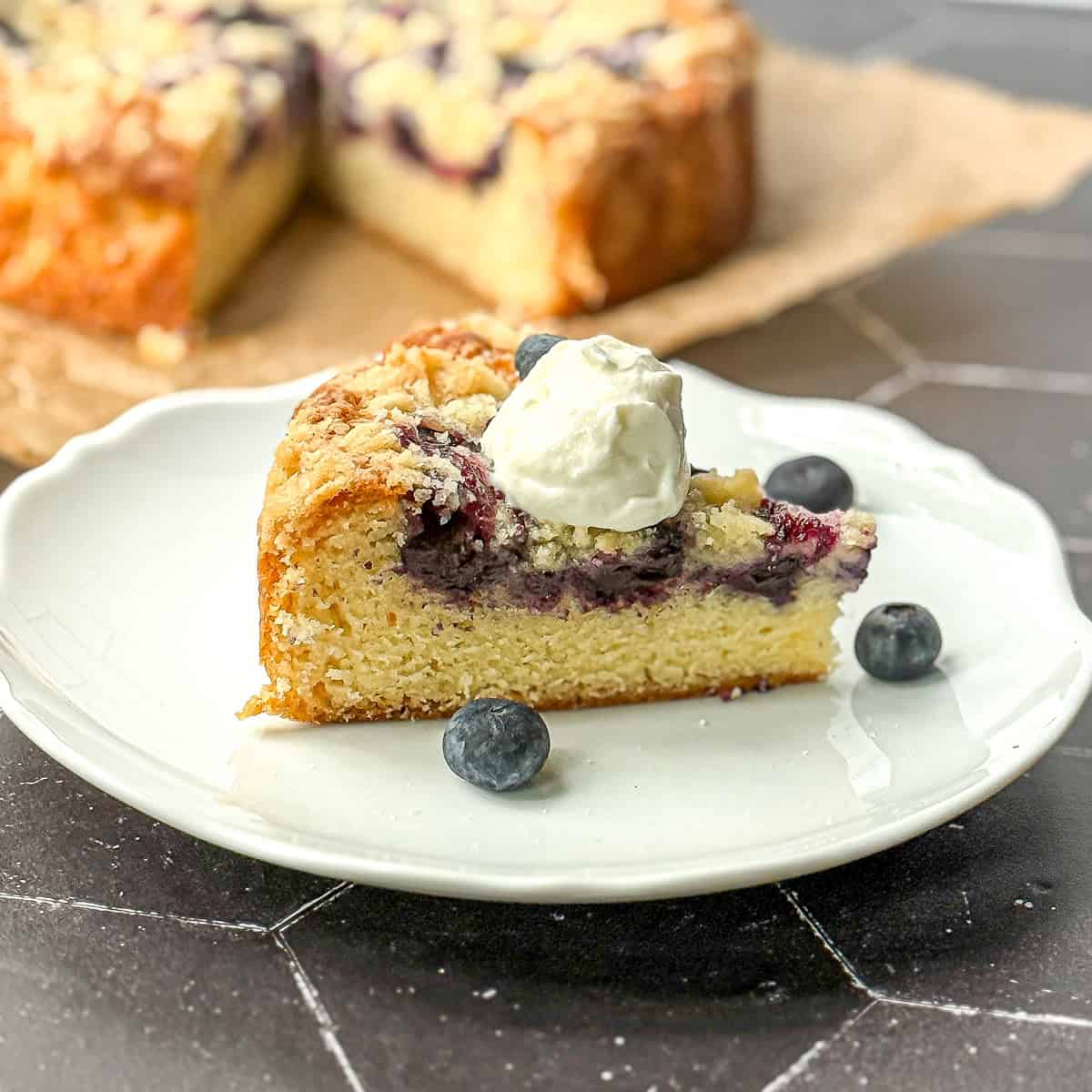 A slice of yogurt cake with blueberries on a white plate with the entire cake behind it on a black background.