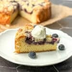 A slice of yogurt cake with blueberries on a white plate with the entire cake behind it on a black background.