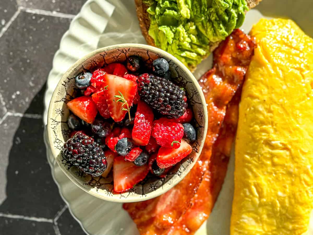 A bowl of mixed berry fruit salad on a plate with avocado toast, bacon, and an omelette.