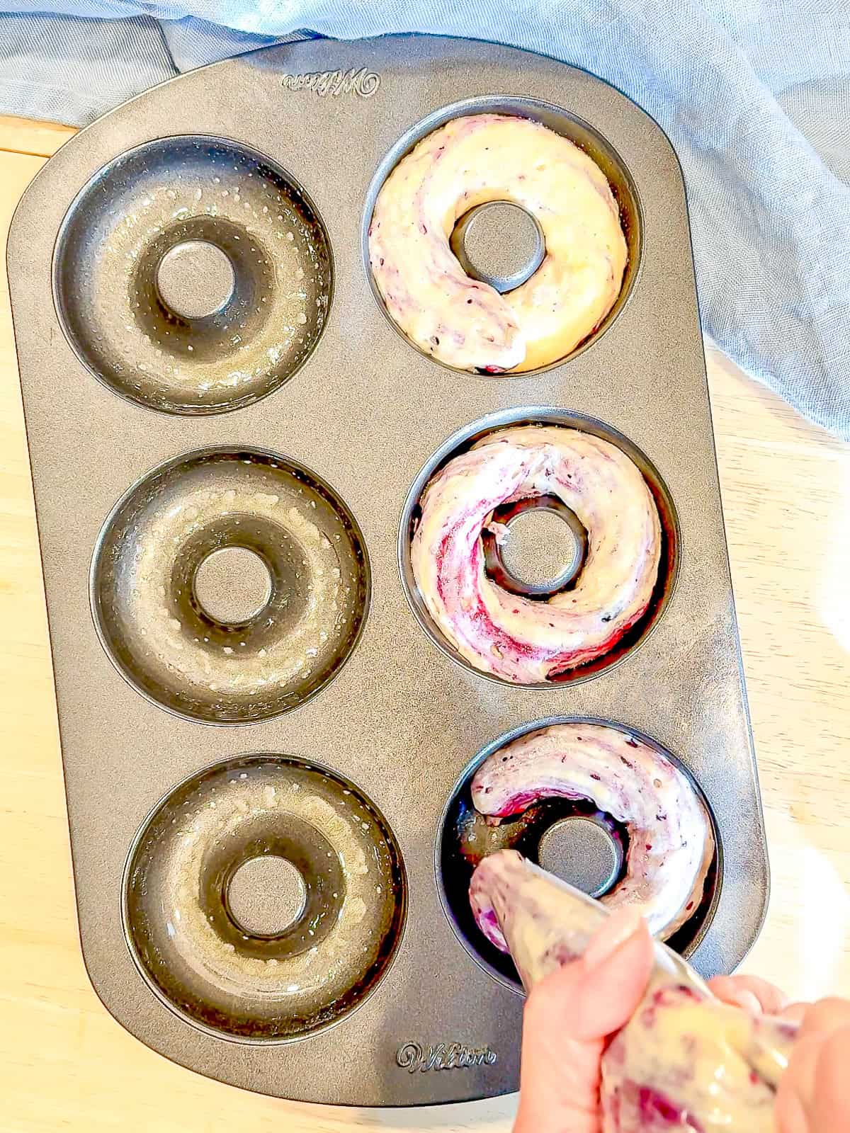 Piping blueberry swirl donut batter into a donut pan.