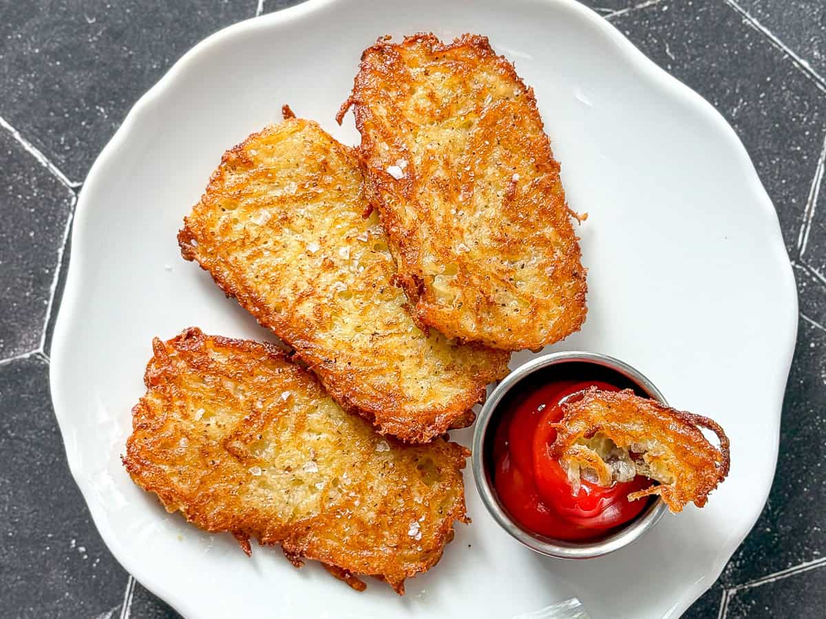 Three hash brown patties on a white plate with a cup of ketchup with a small piece of hash brown in it.