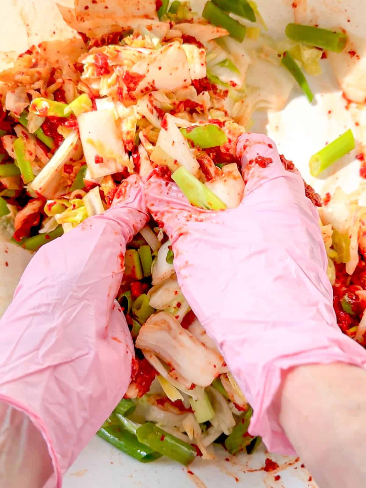 Glove hands mixing kimchi paste into cabbage and green onion in a pink bowl.