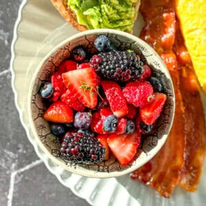 A bowl of mixed berry fruit salad on a plate with avocado toast, bacon, and an omelette.