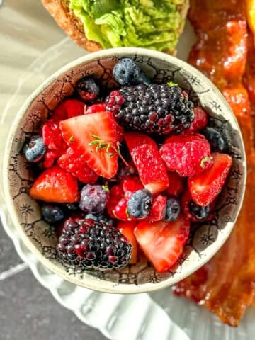 A bowl of mixed berry fruit salad on a plate with avocado toast, bacon, and an omelette.