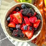 A bowl of mixed berry fruit salad on a plate with avocado toast, bacon, and an omelette.