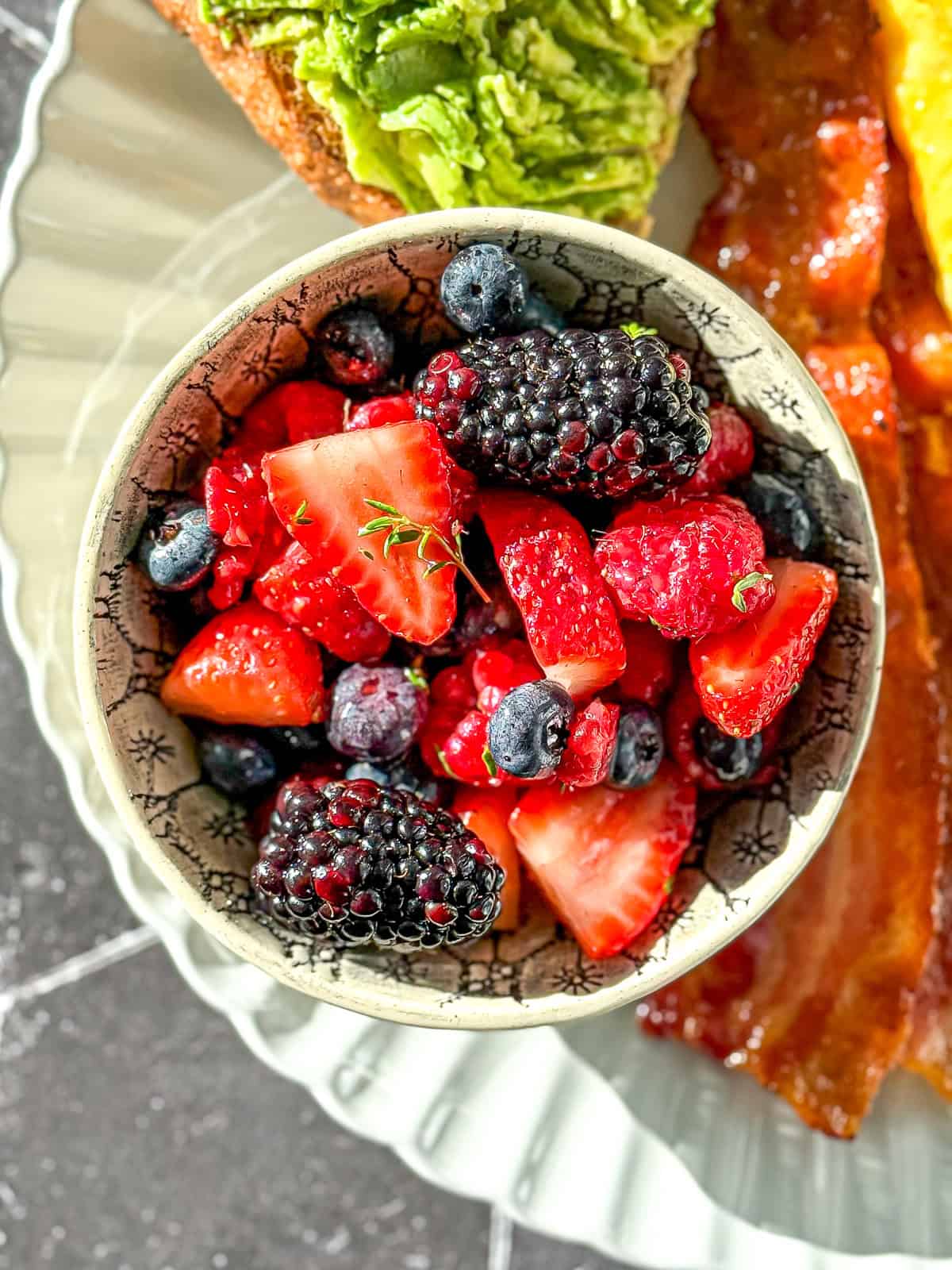 A bowl of mixed berry fruit salad on a plate with avocado toast, bacon, and an omelette.