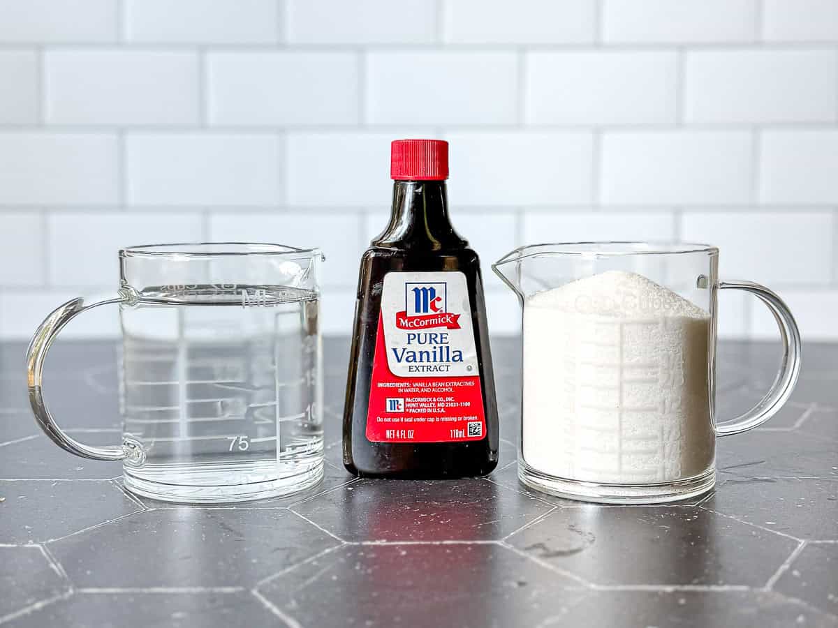 Two measuring cups containing water and granulated sugar and a bottle of vanilla extract.