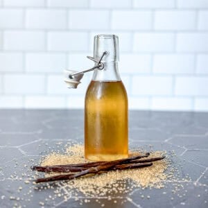 Pinterest image of a bottle of vanilla simple syrup on a black counter surrounded by sugar and vanilla bean pods.