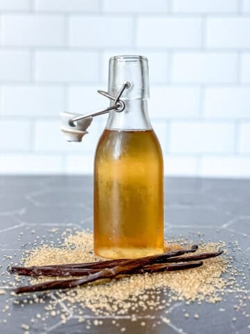 Pinterest image of a bottle of vanilla simple syrup on a black counter surrounded by sugar and vanilla bean pods.
