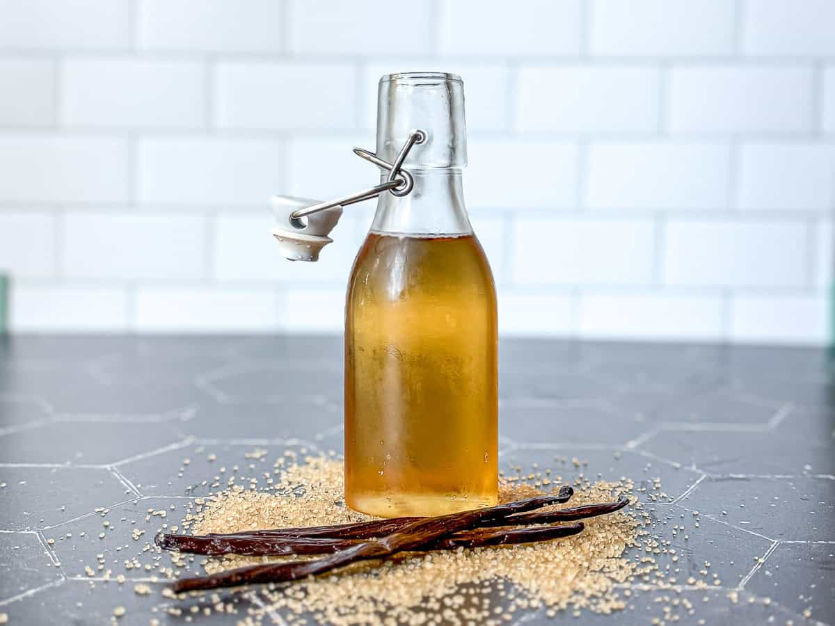 Pinterest image of a bottle of vanilla simple syrup on a black counter surrounded by sugar and vanilla bean pods.
