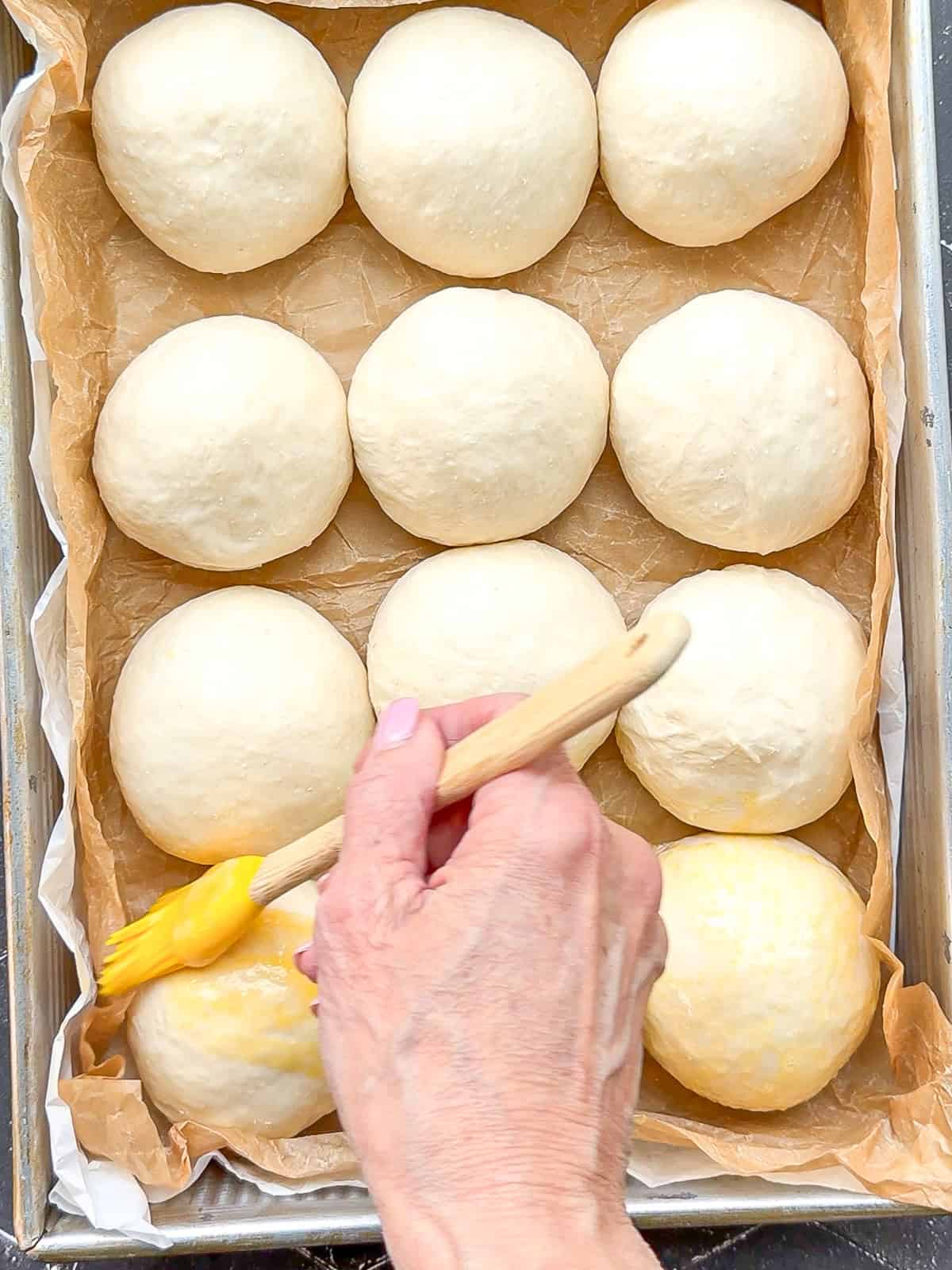 Brushing unbagged dinner rolls with an egg wash in a parchment lined pan.