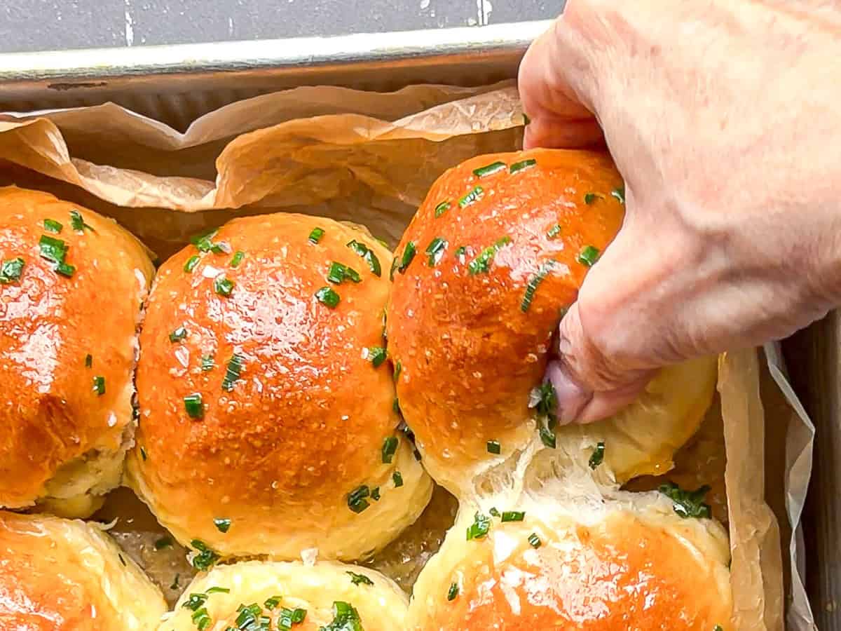 Hand pulling a warm dinner roll out of a pan lined with parchment paper.