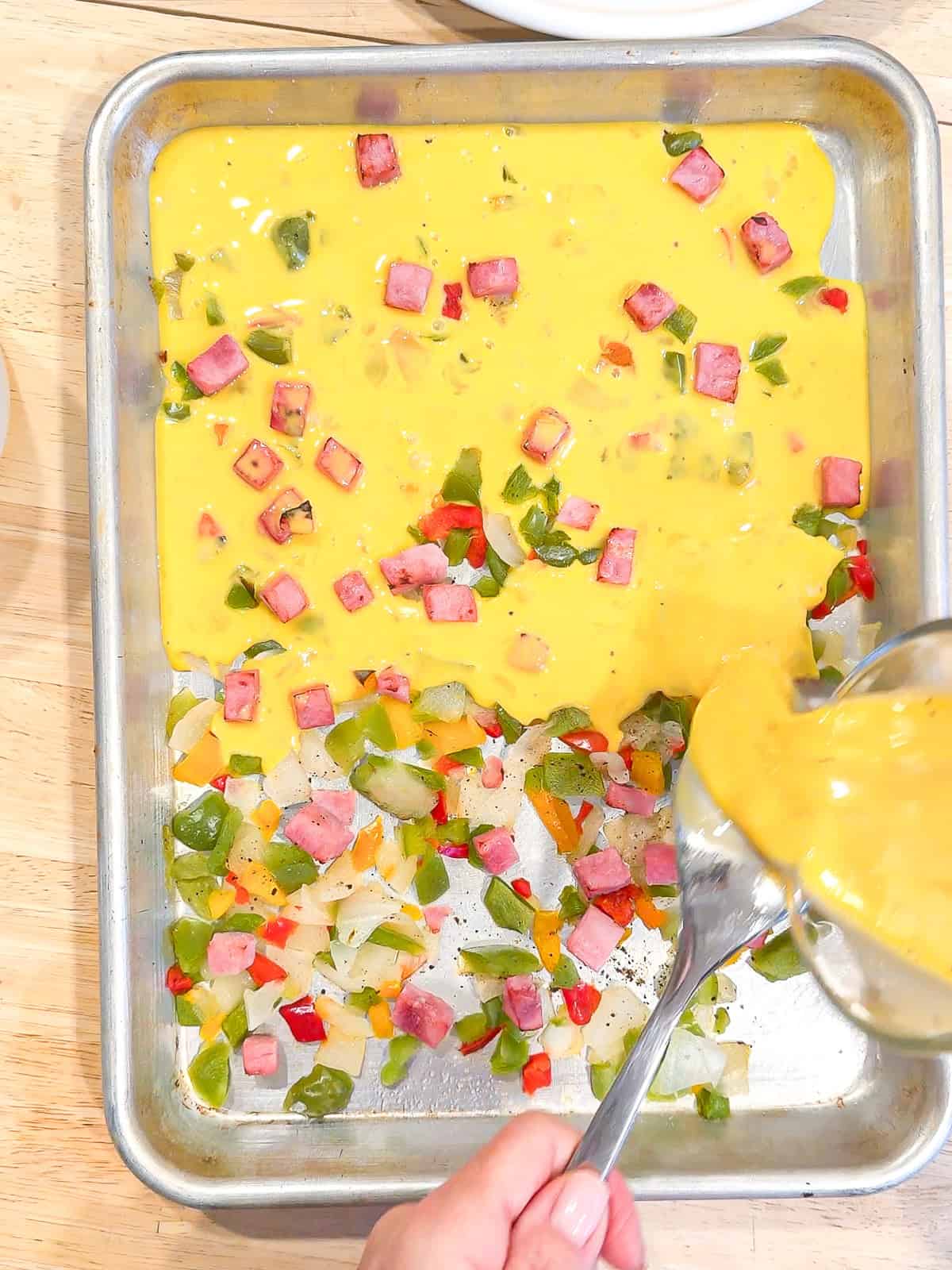 Pouring eggs into a small sheet pan filled with peppers, onions, and ham.