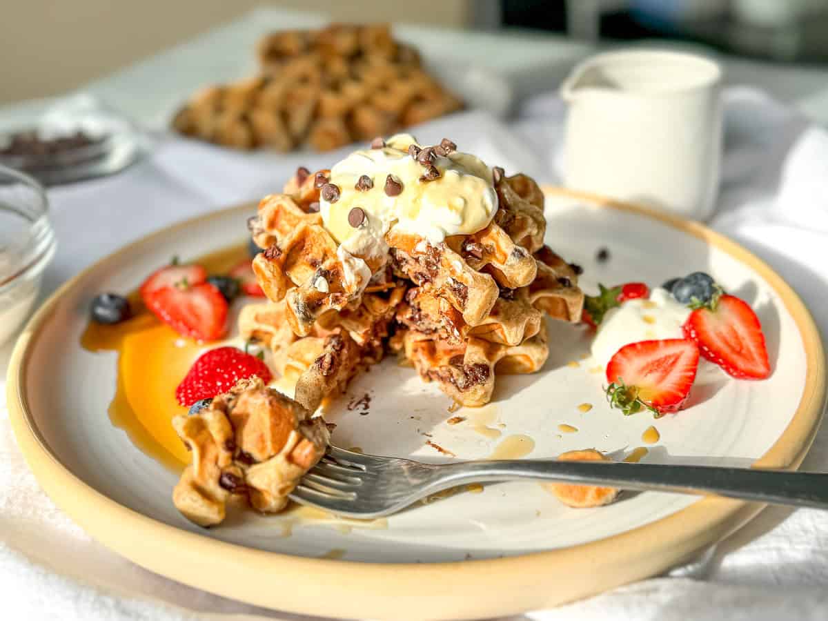 Image of a stack of banana waffles with whipped cream and chocolate chips and fresh fruit on a white and tan plate.