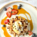 Image of a stack of banana waffles with whipped cream and chocolate chips and fresh fruit on a white and tan plate.