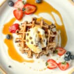 Image of a stack of banana waffles with whipped cream and chocolate chips and fresh fruit on a white and tan plate.