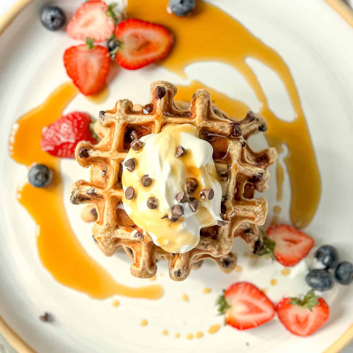 Image of a stack of banana waffles with whipped cream and chocolate chips and fresh fruit on a white and tan plate.