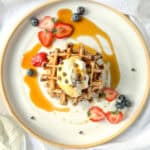 Image of a stack of banana waffles with whipped cream and chocolate chips and fresh fruit on a white and tan plate.