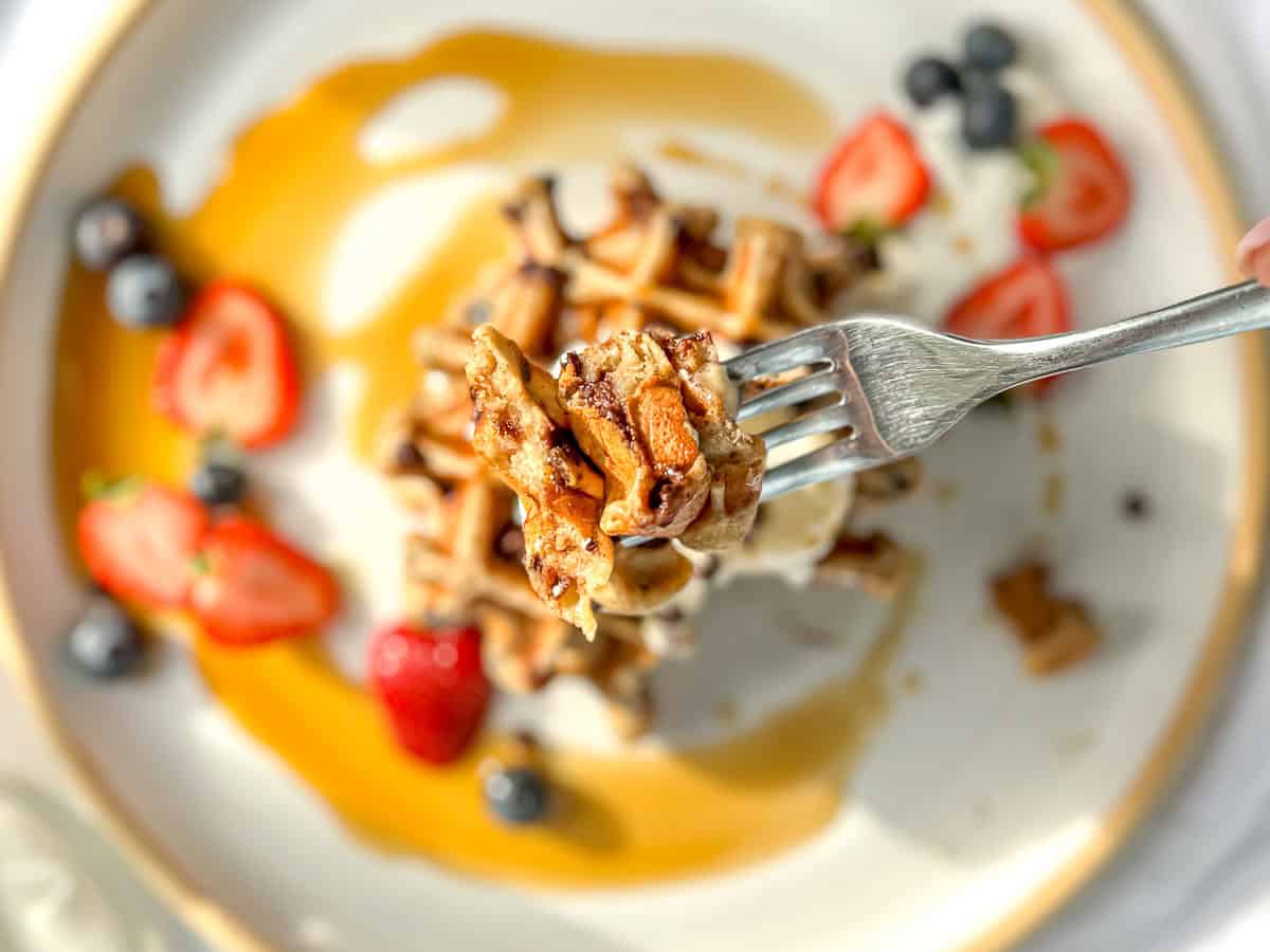 A fork holding a large bite of banana and chocolate chip waffles over a plate of waffles.