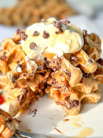 Image of a stack of banana waffles with whipped cream and chocolate chips and fresh fruit on a white and tan plate.