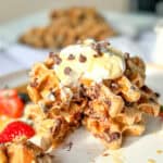 Image of a stack of banana waffles with whipped cream and chocolate chips and fresh fruit on a white and tan plate.