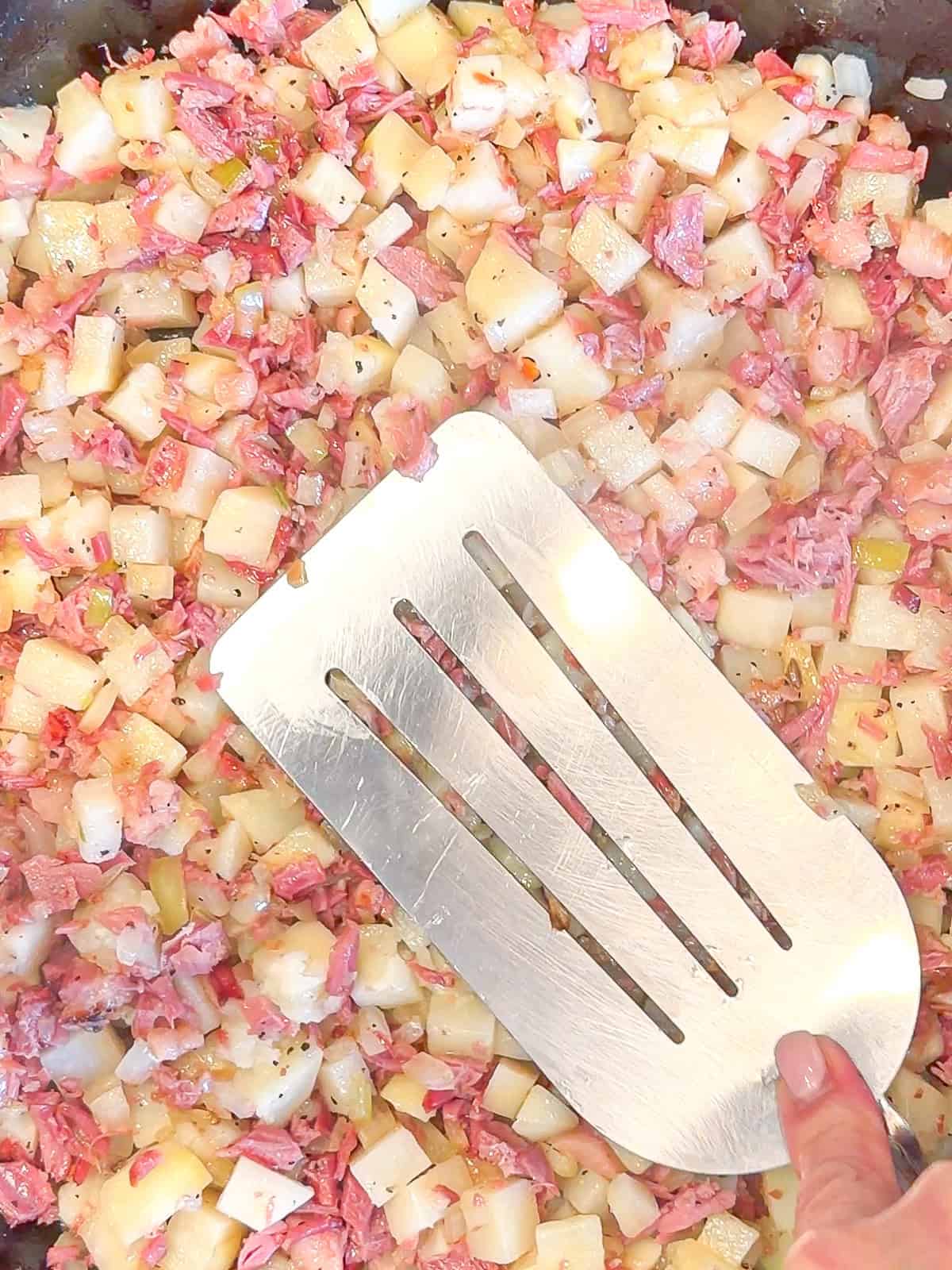 Using a large spatula to press corn beef and potatoes into a hot cast-iron pan.