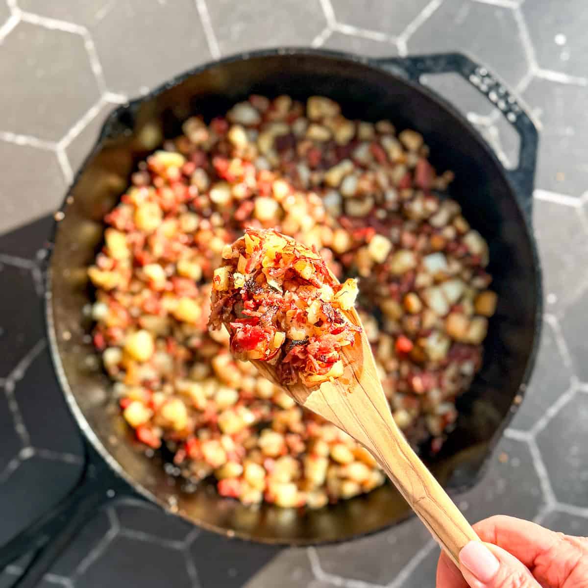 Wooden spoon holding corned beef hash on top of a cast-iron skillet filled with more hash.