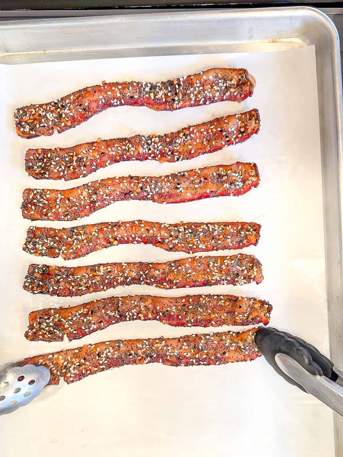 Using tongs to place bacon strips on a parchment paper lined baking sheet.