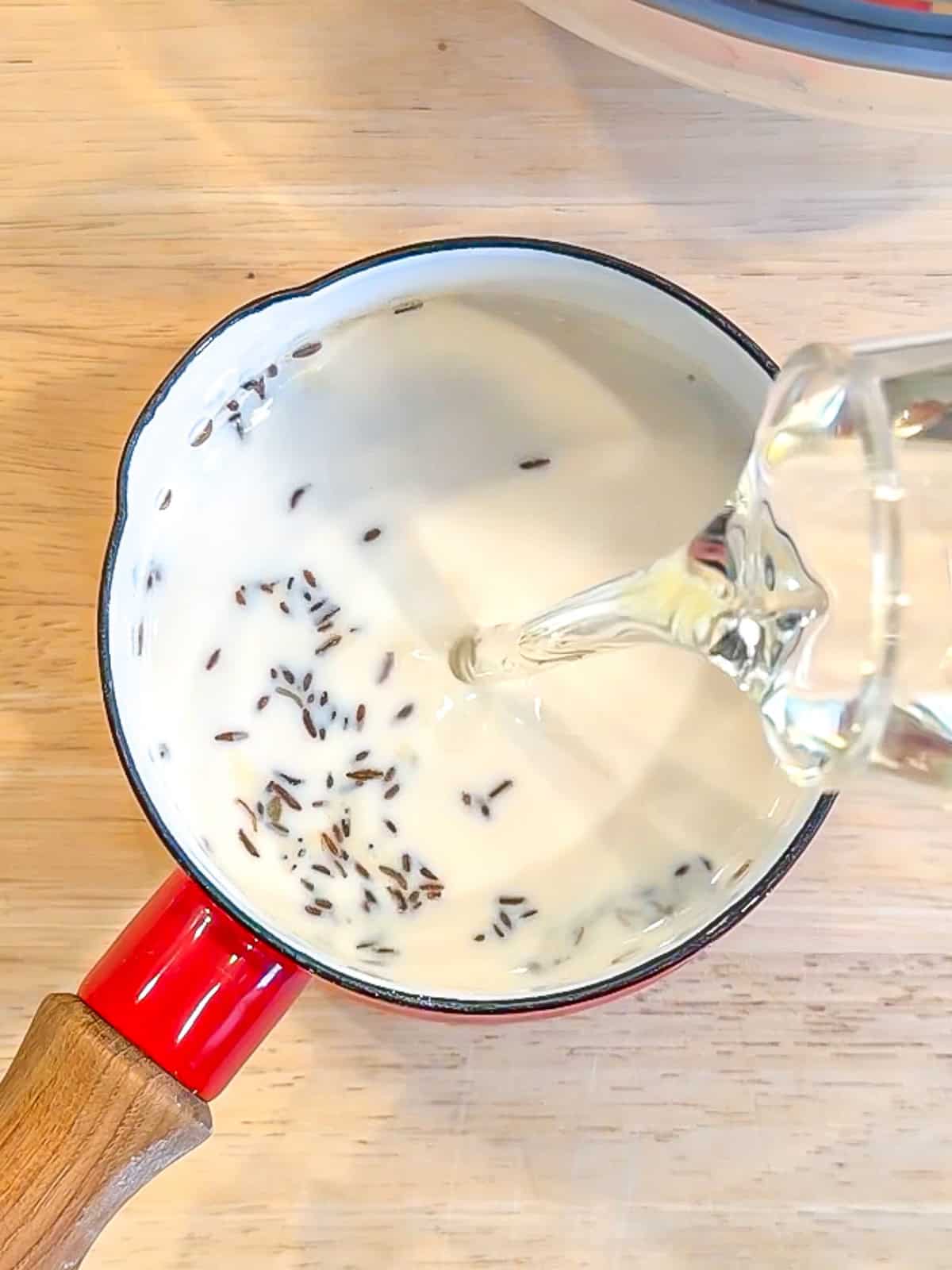 Adding oil to milk, water, and caraway seeds in a red sauce pan.