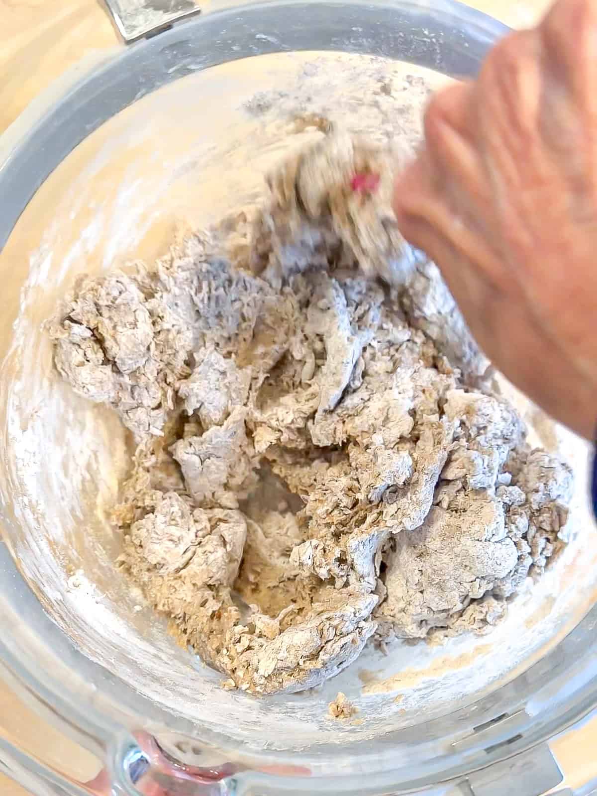 Hand mixing wet and dry ingredients to make rye bread in a glass bowl.