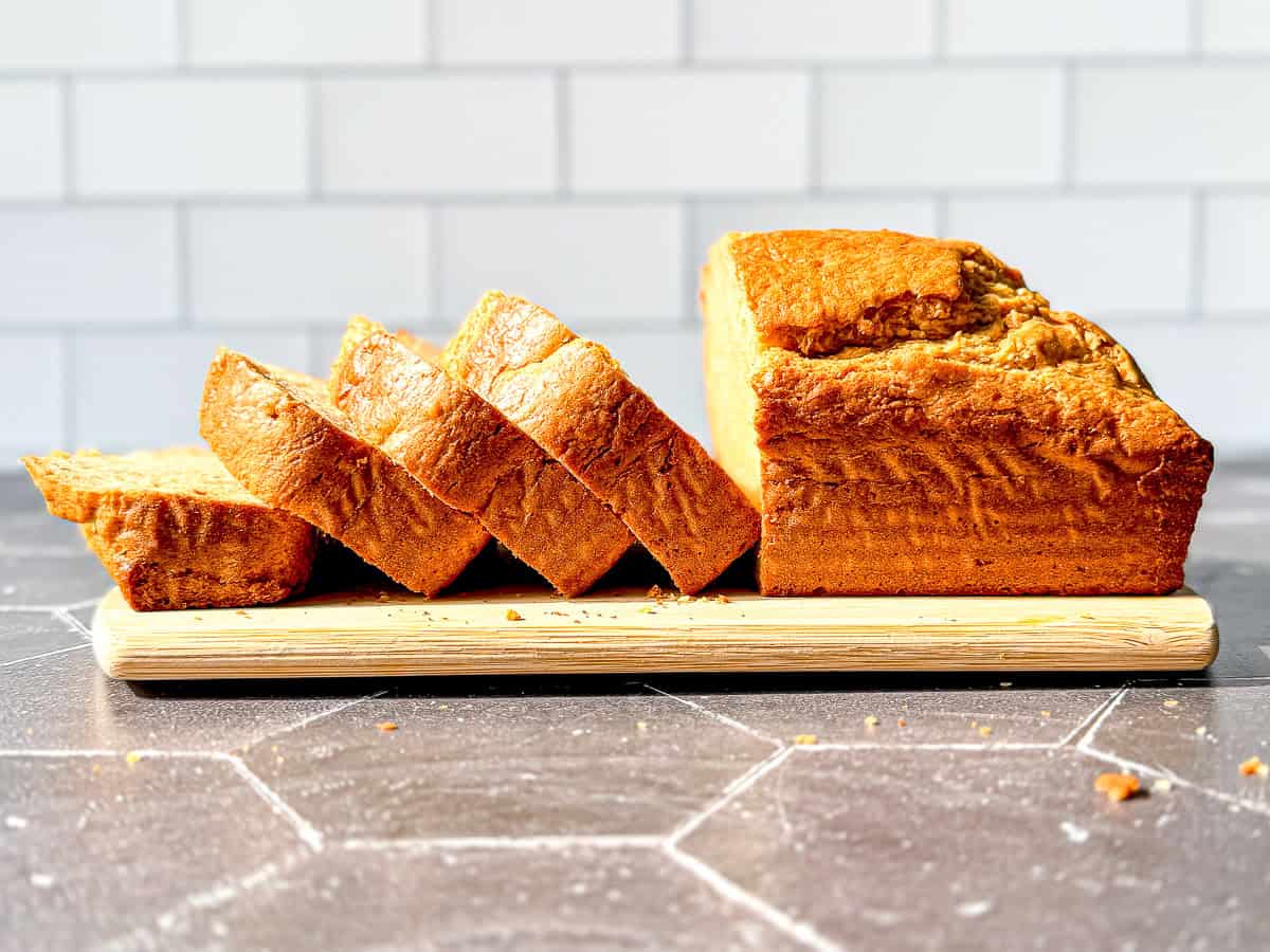 A sliced loaf of banana bread on a wooden cutting board in front of a tile backdrop.