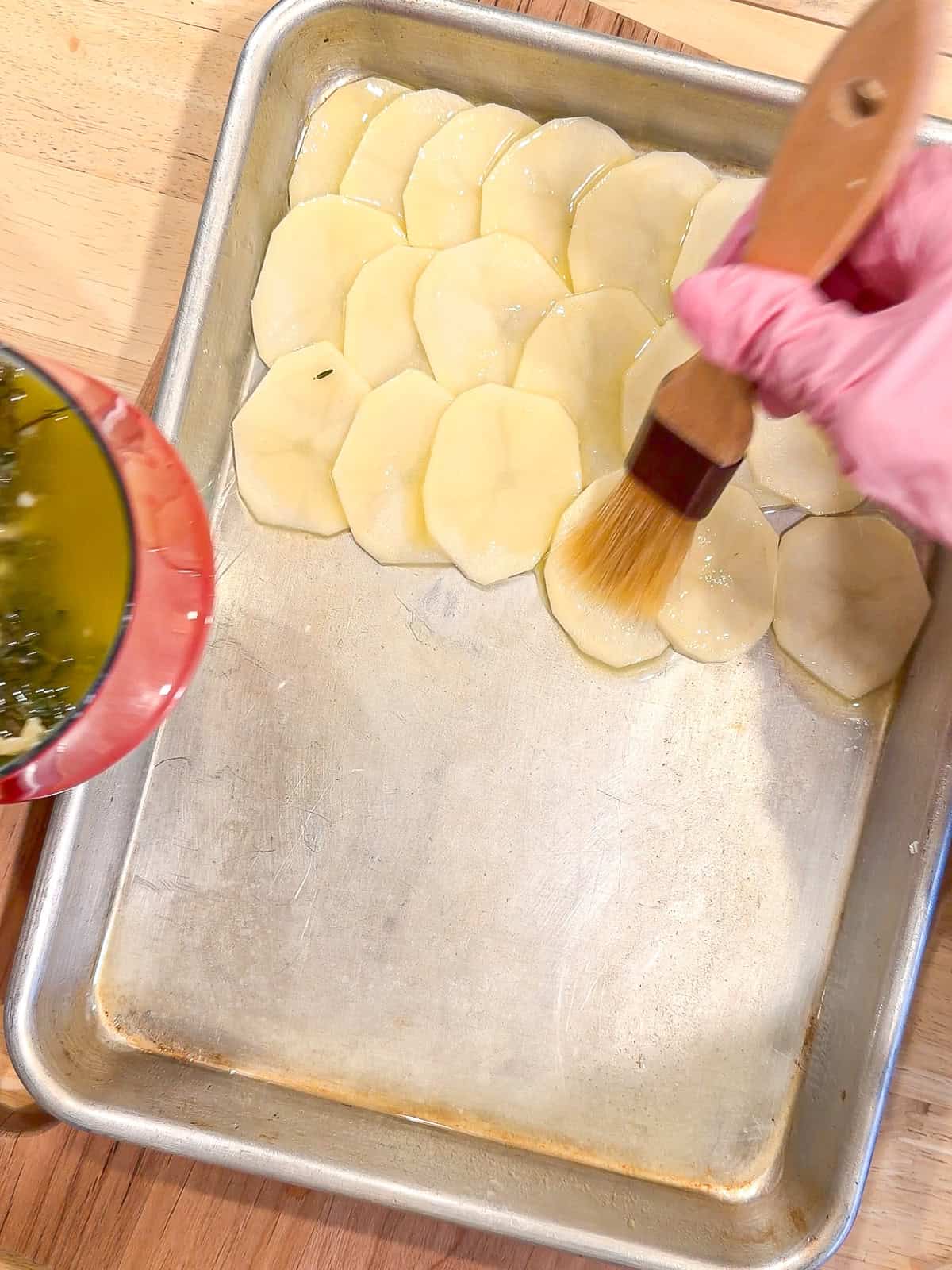 Brushing melted butter on top of sliced potatoes in a sheet pan.