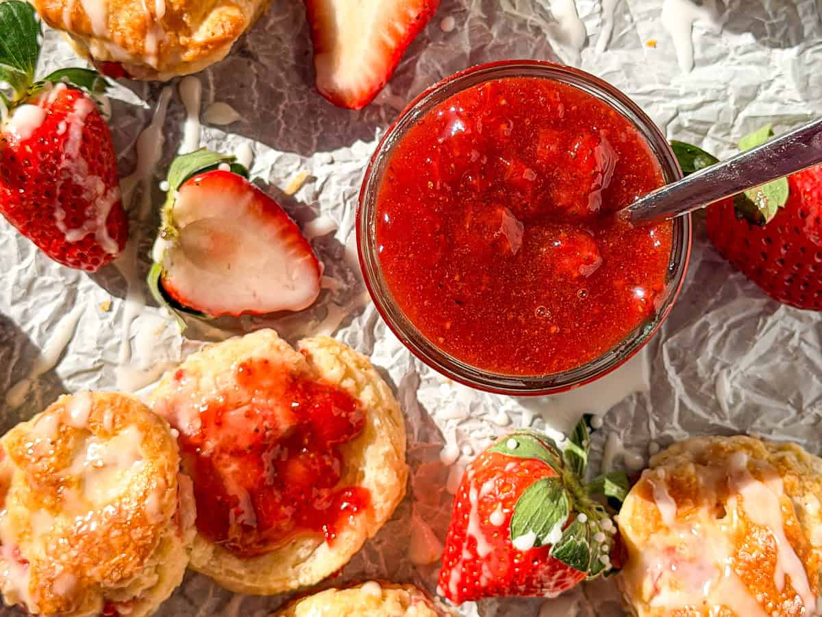 A jar of strawberry jam with a spoon in it surrounded by strawberry biscuits and fresh strawberries.