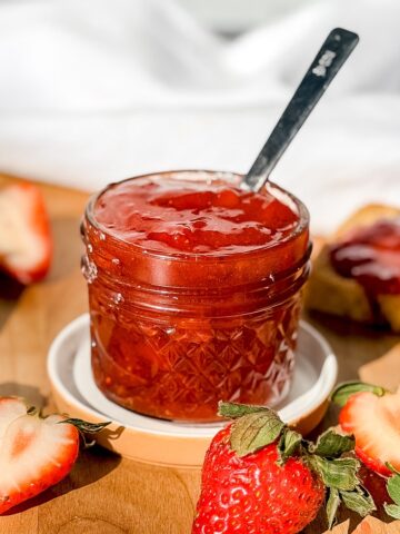 A jar of strawberry jam with strawberries and a piece of toast in the background.